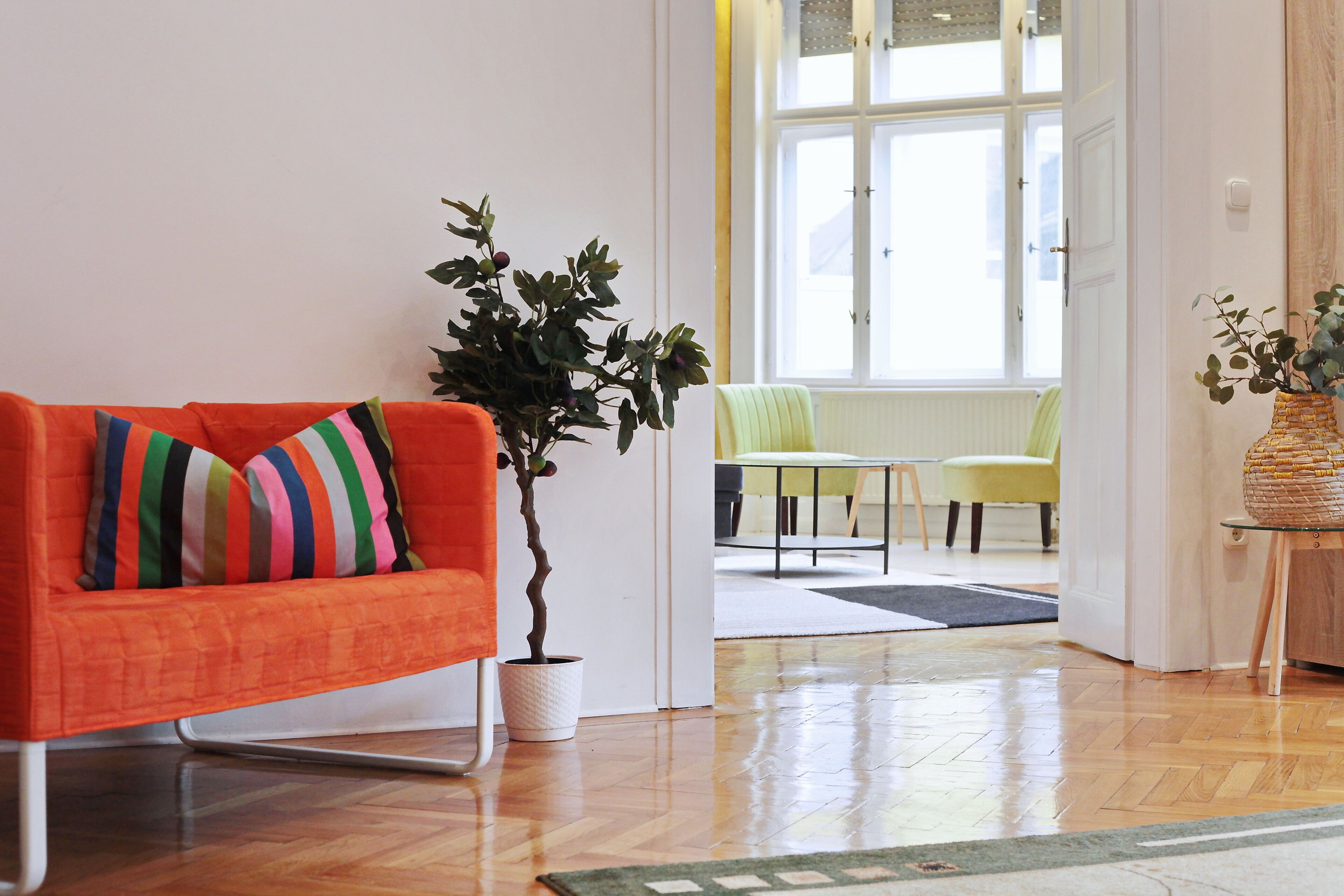 Una sala de estar moderna con un sofá naranja brillante, una planta en maceta y vista a un comedor a través de una puerta abierta.