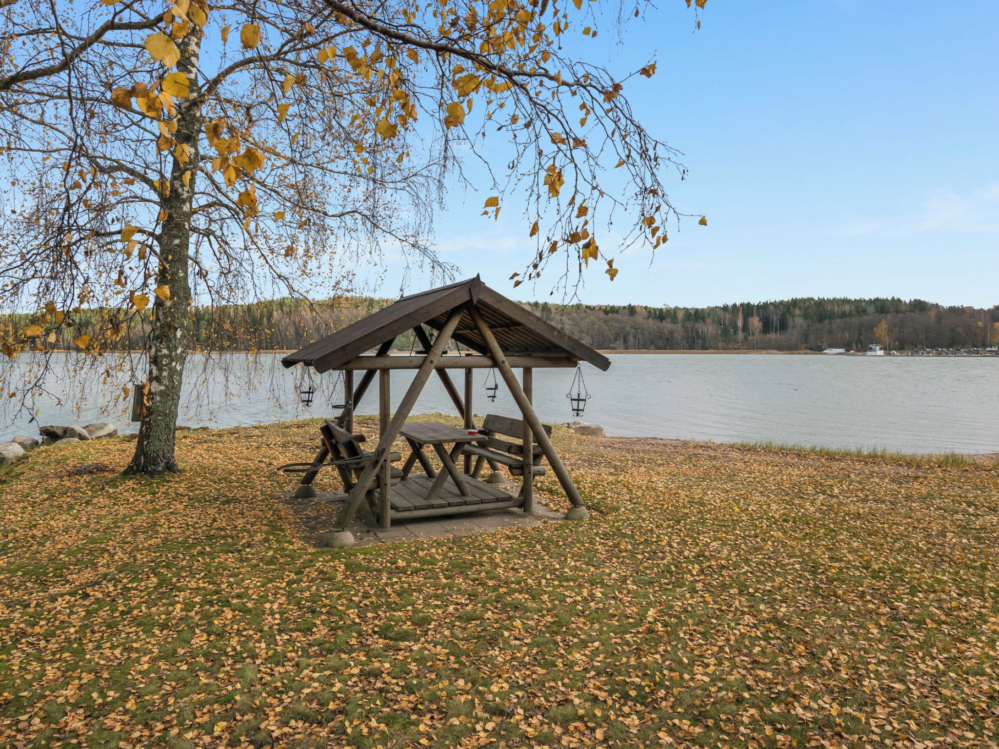 En träbeklädd picknickplats vid en sjö, med bänkar och en gunga, omgiven av höstlöv.