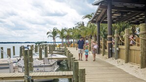 A wooden pier leading to a dock with boats, palm trees, and a covered area with seating.