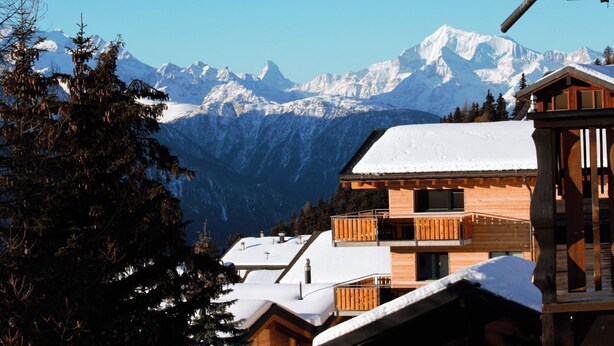 A wooden chalet with a balcony, surrounded by snow-covered mountains and trees.