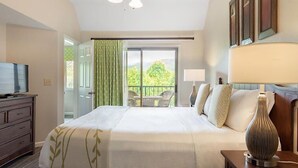Ein Hotelzimmer mit einem Bett, einem Fernseher, einer Lampe, einem Schminktisch und einem Balkon mit Blick auf Grünflächen.