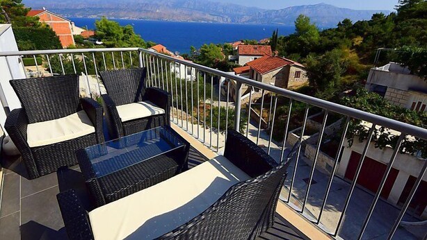 A balcony with wicker furniture and a glass table overlooking a coastal view with houses and mountains in the distance. - Molo Lozna by Interhome (Brač/Postira)