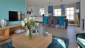 Uma sala de estar moderna com televisão, uma mesa de centro com um vaso de flores e um espaço de jantar com cadeiras e mesa.