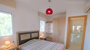 Un dormitorio con cama, mesitas de noche, una lámpara colgante roja, una ventana y un baño visible a través de una puerta abierta.