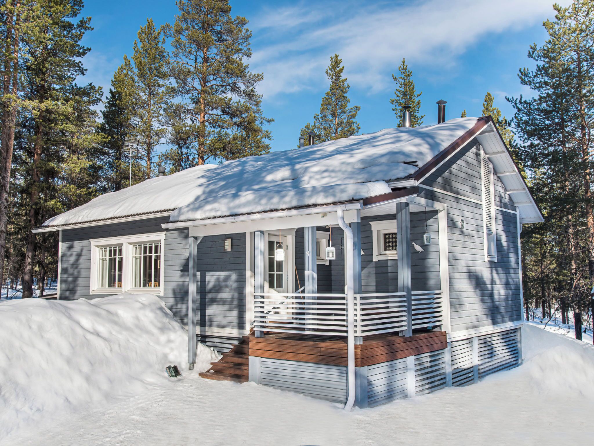 A small, single-story cabin with a porch, surrounded by snow and tall pine trees.