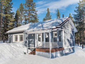 Une petite cabane à un seul étage, dotée d’un porche, entourée de neige et de grands pins.
