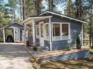 Une petite cabane avec un porche, un barbecue et un abri de jardin, située dans une zone boisée.