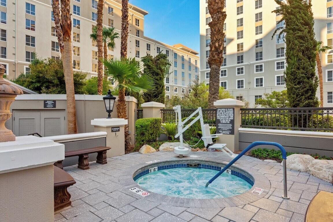 A hot tub surrounded by stone benches, palm trees, and a poolside area with a view of a high-rise building.