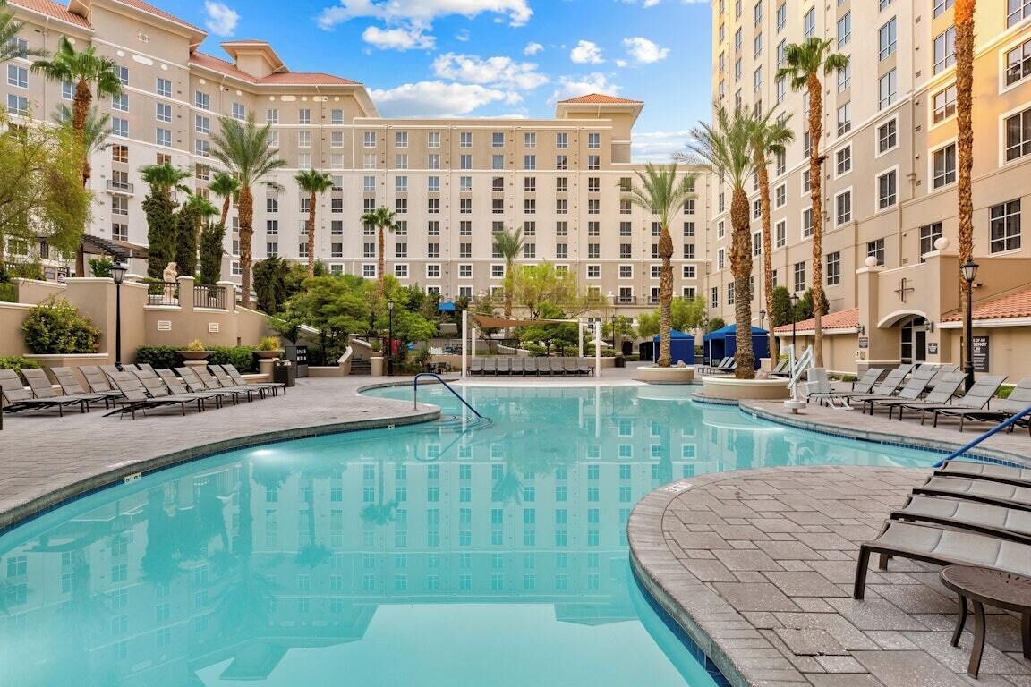 A hotel pool area with lounge chairs, palm trees, and a large building in the background.