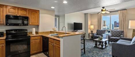 A modern kitchen with wooden cabinets, a black stove, and a microwave. There is a dining area with a table and chairs, and a living room with a sofa and a TV.