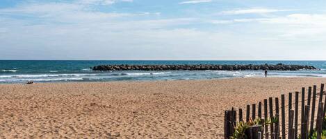 A sandy beach with a wooden fence and a rocky breakwater in the distance.