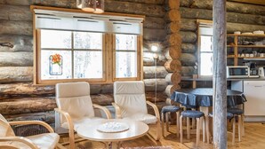 A cozy log cabin interior with a dining area, two white leather chairs, a round table, and a kitchen area with a microwave and coffee maker.