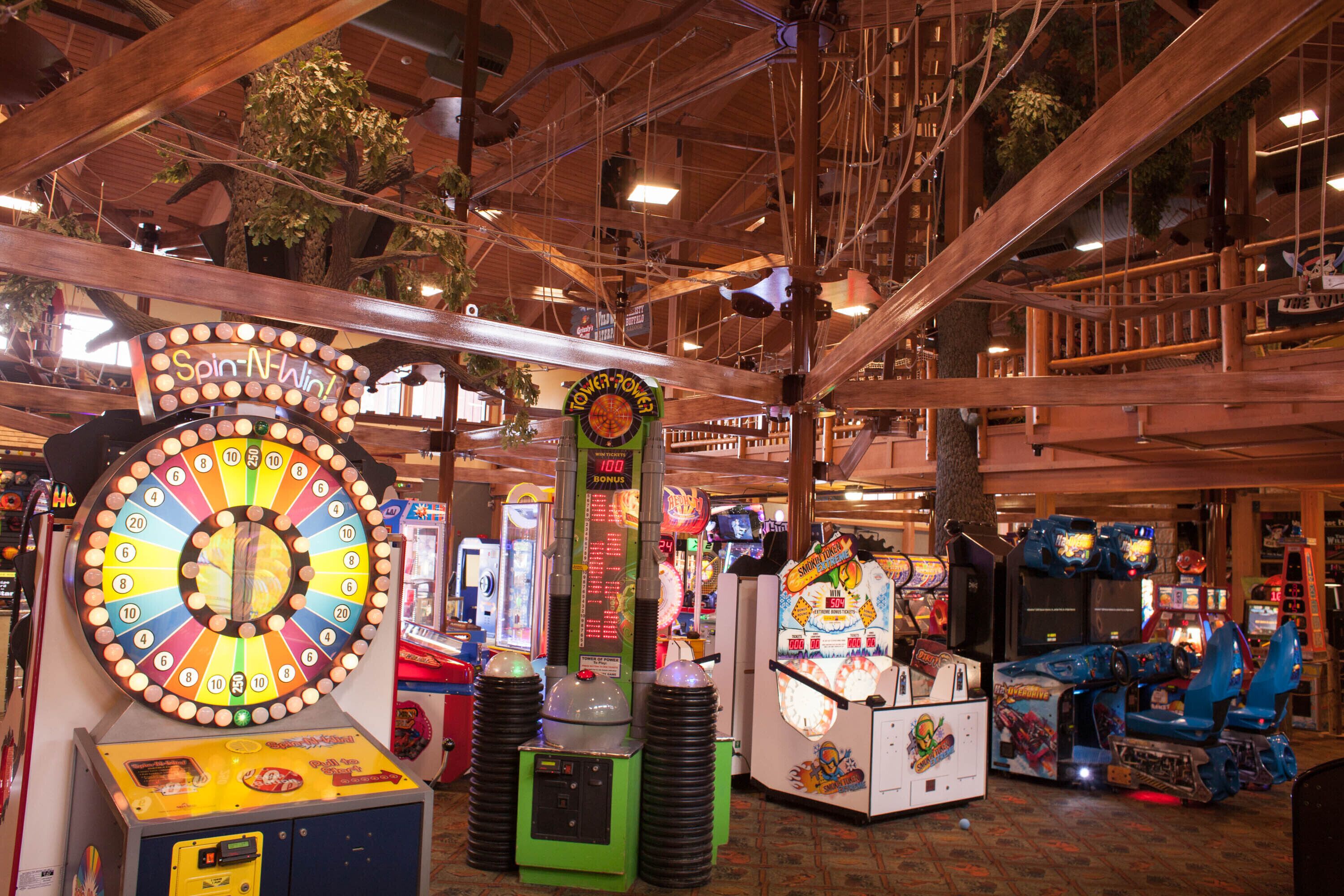 Variedad de juegos de arcade y una ruleta en un espacio interior con vigas de madera y plantas artificiales.