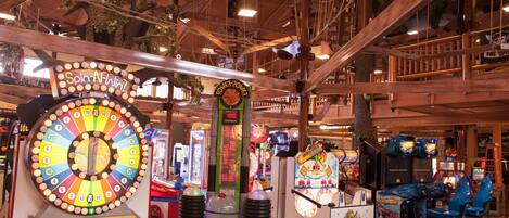 Variedad de juegos de arcade y una ruleta en un espacio interior con vigas de madera y plantas artificiales.