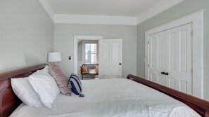 Ein Hotelzimmer mit einem großen Bett, einem hölzernen Kopfteil, einem Nachttisch mit einer Lampe und Blick auf den Flur sowie ein weiteres Zimmer.