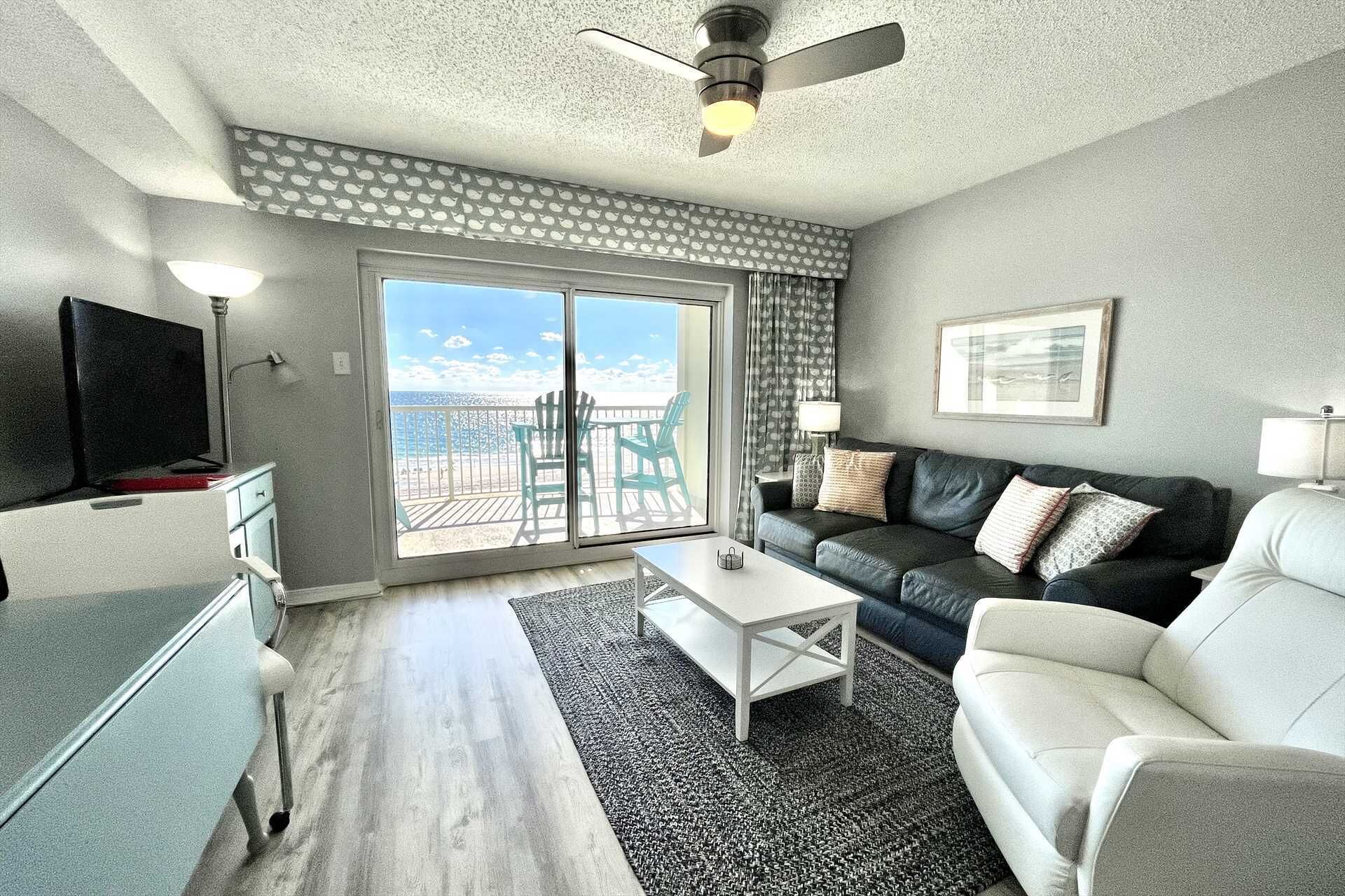 Un salon avec un canapé, une table basse et un téléviseur. Il y a un balcon avec vue sur la plage.
