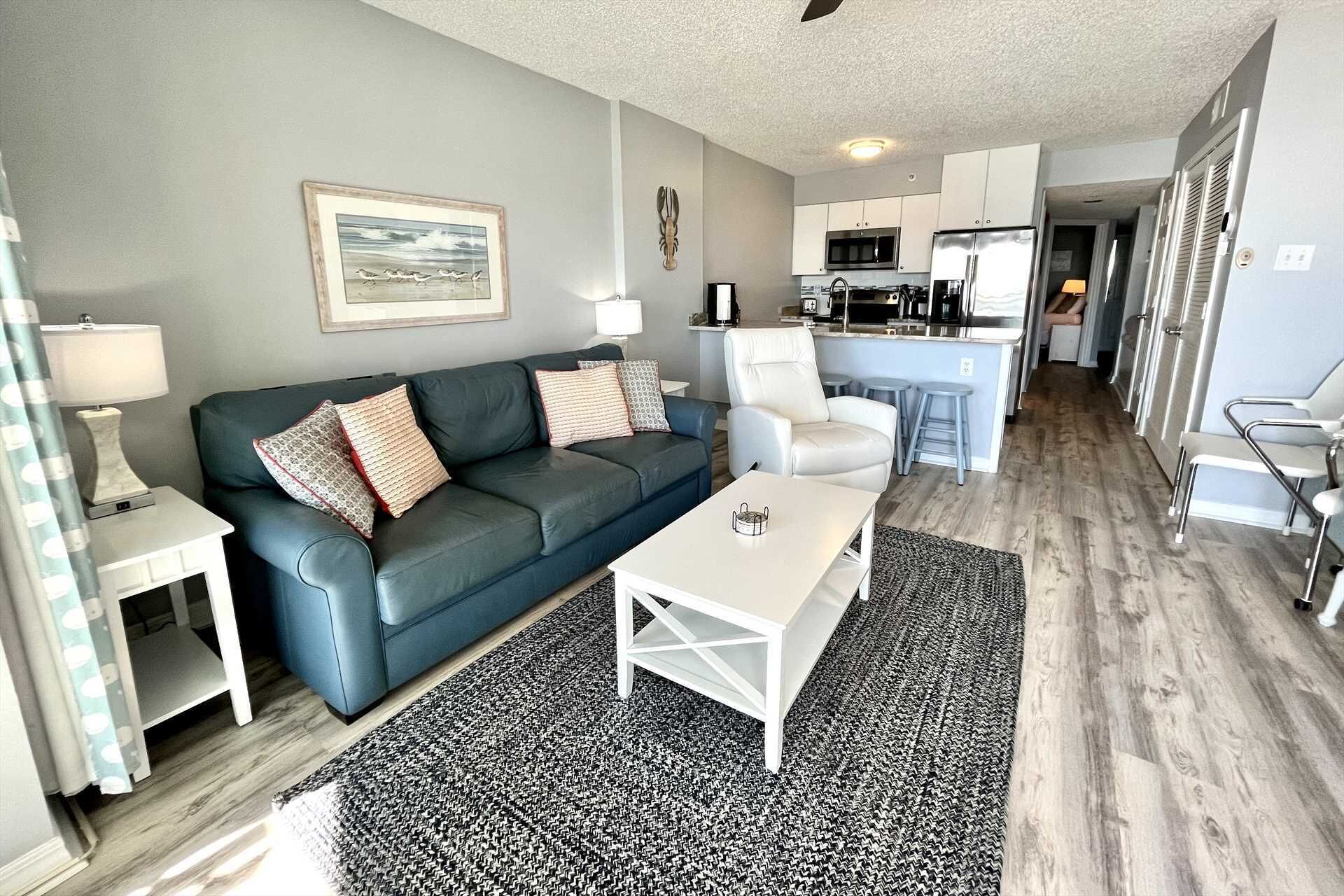 A modern living room with a dark blue sofa, white coffee table, and patterned rug. A kitchen with white cabinets and stainless steel appliances is visible in the background.