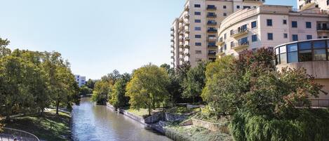 A riverside walkway with a bridge, surrounded by trees and modern buildings.