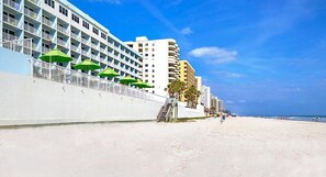 Un hotel frente a la playa con balcones, una playa de arena y un cielo azul despejado.