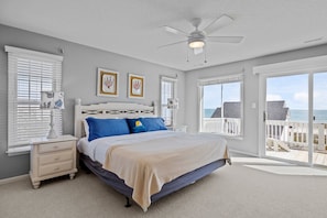 A bedroom with a bed, a nightstand, a ceiling fan, and a view of the beach through the door.