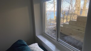 A corner of a room with a bed against a wall, a window with a view of a wooden deck and trees.