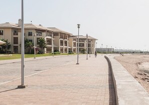 Ein gepflasterter Weg führt zu einem Gebäude mit mehreren Balkonen, eine Sandküste rechts daneben und Straßenlaternen säumen den Weg.