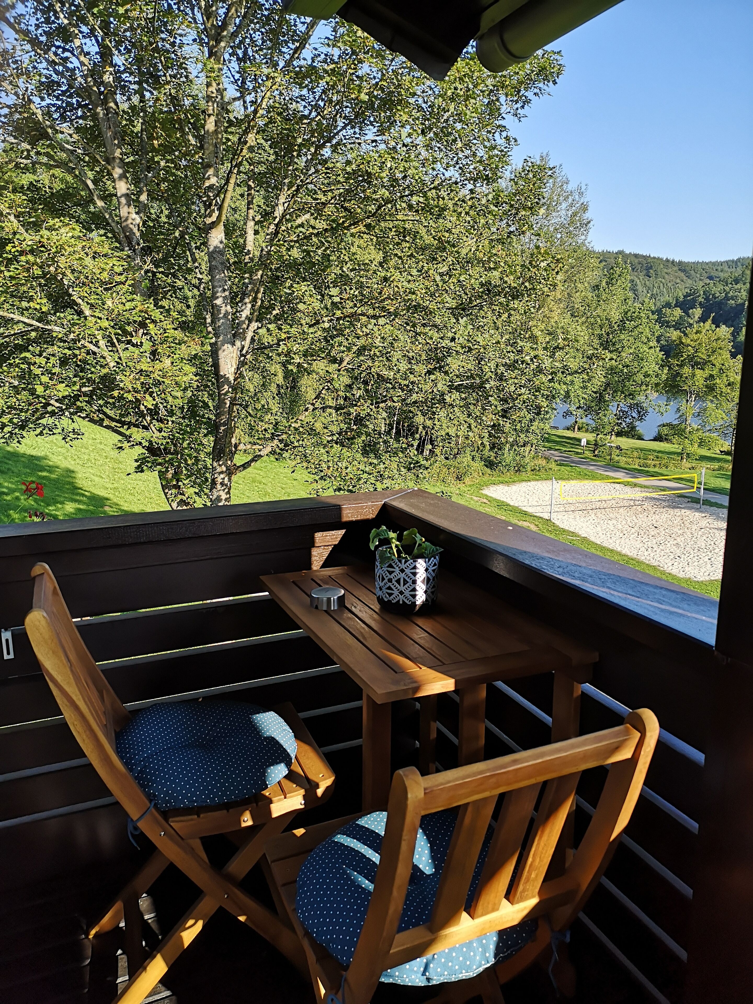 A wooden deck with a table and two chairs, overlooking a lake and forested hills.
