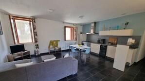 Una sala de estar moderna con un sofá gris, una mesa de comedor con sillas, una zona de cocina con armarios blancos y un televisor sobre un mueble de madera.