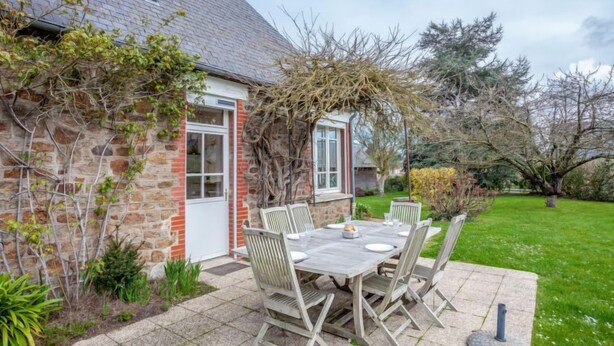 Een stenen gebouw met een betegeld terras, een houten tafel met stoelen, en een tuin vol planten.
