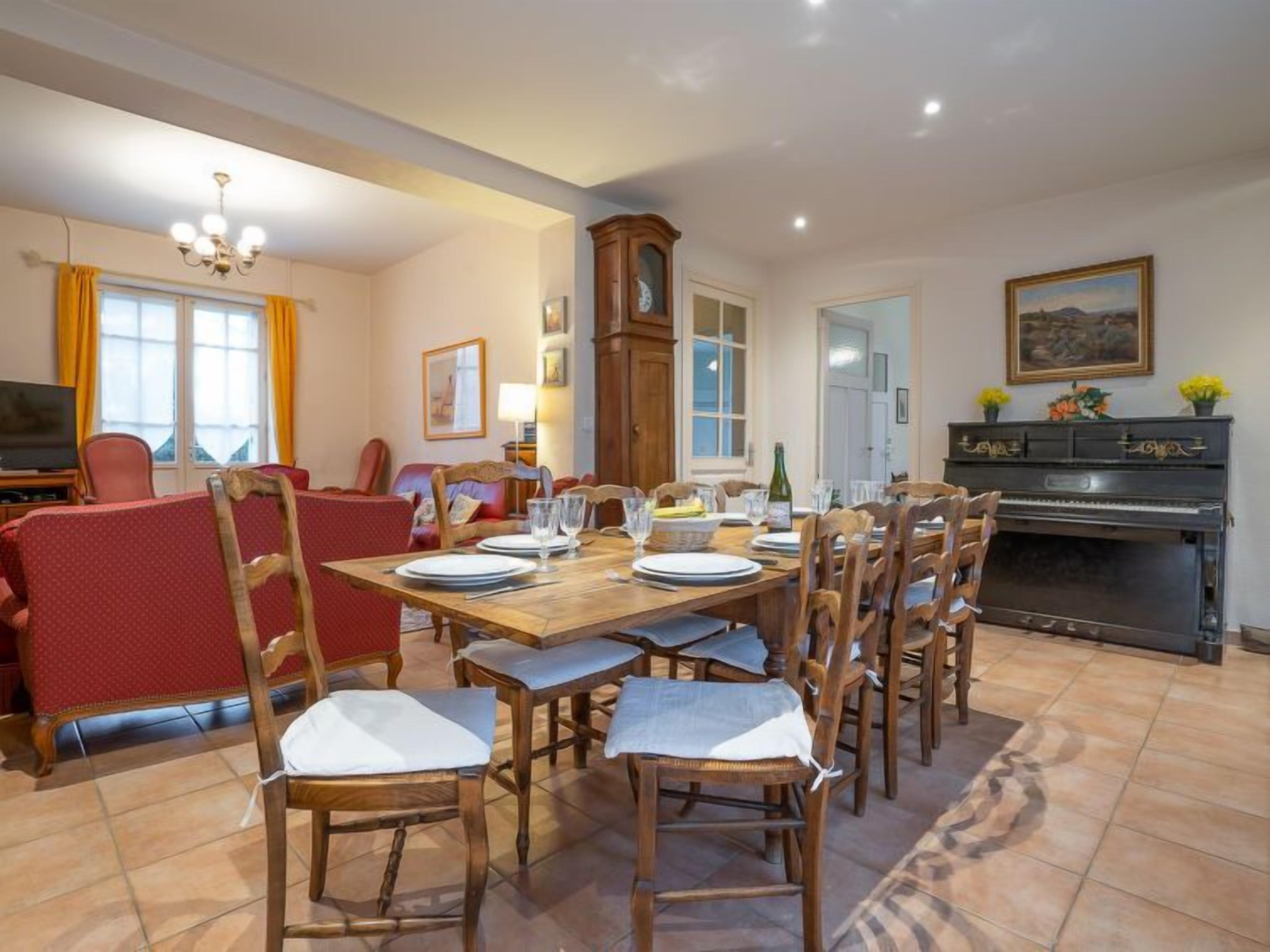 A dining area with a wooden table set for six, surrounded by wooden chairs. A piano is against the wall, and a television is on a stand to the left.