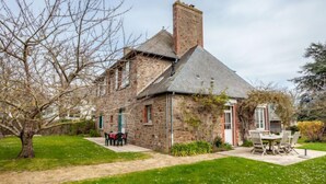 Een stenen huis met schoorsteen, een terras met buitenmeubilair en een verzorgd gazon.