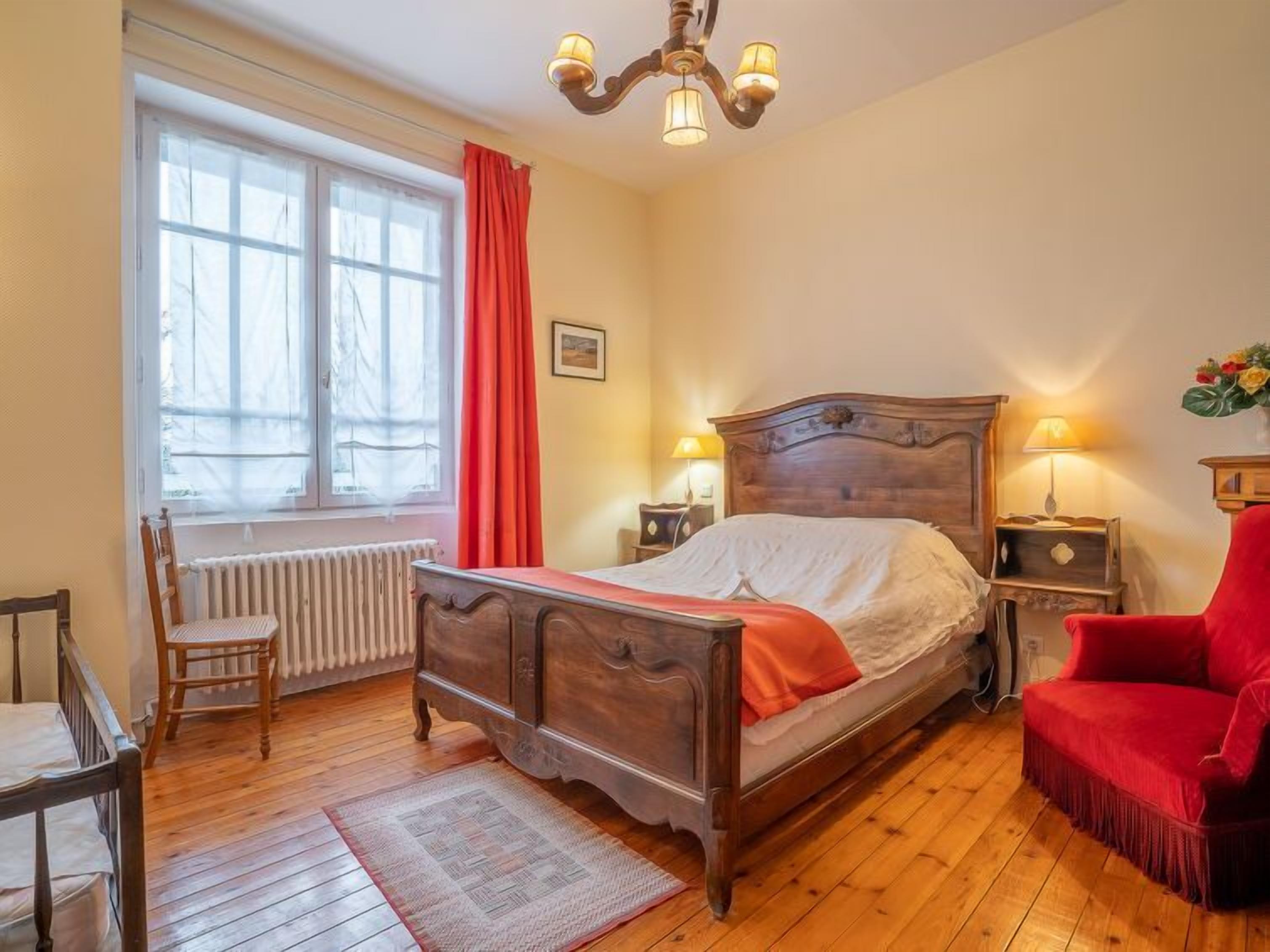 Een slaapkamer met een houten bed, een rode stoel, een radiator, een raam met gordijnen en een kroonluchter.
