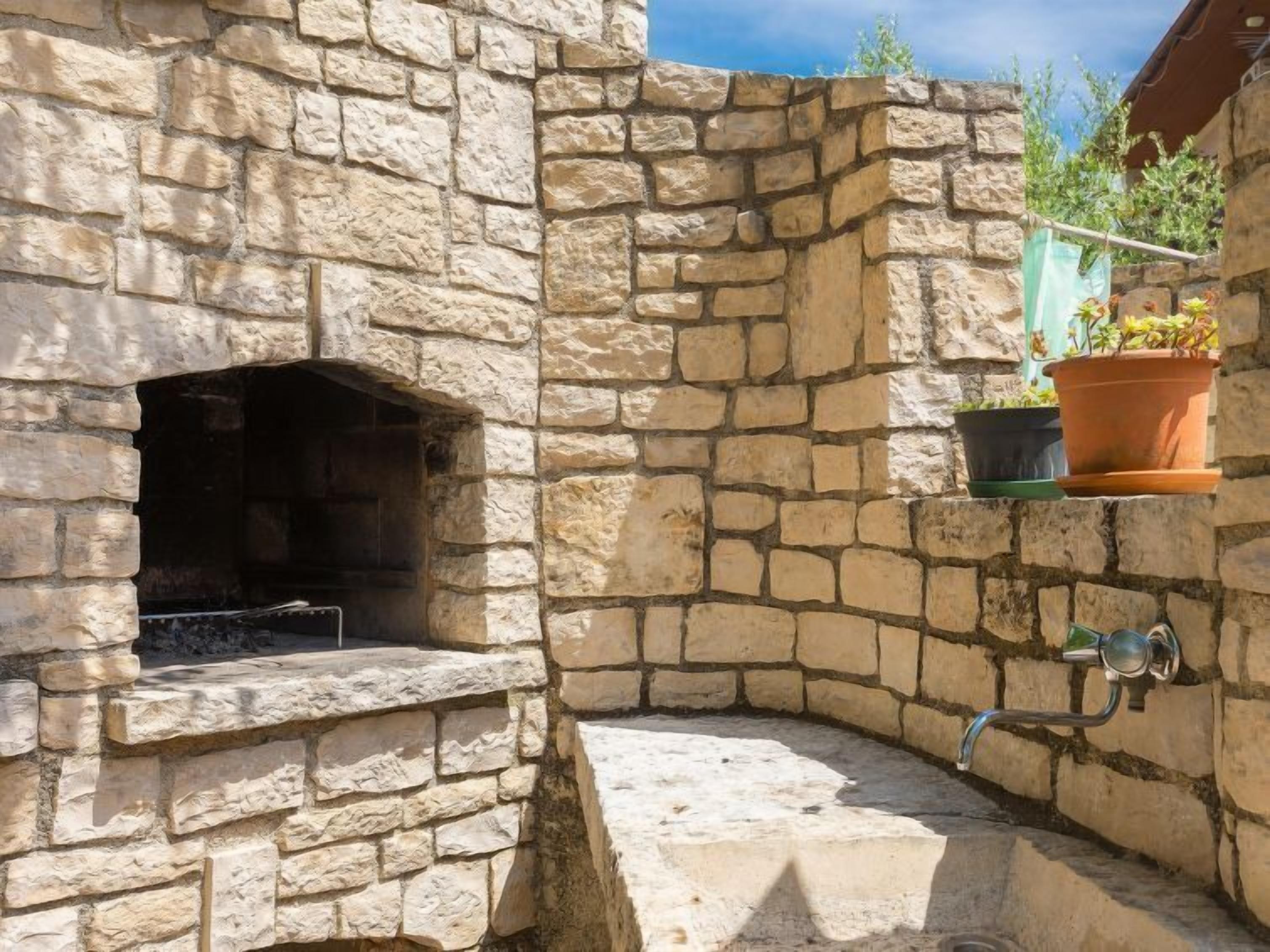 Un espacio al aire libre de piedra con horno de pizza empotrado, una pequeña zona de estar y plantas en macetas.