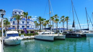 Ein Yachthafen mit mehreren dort vertäuten Yachten, ein Gebäude mit blau-weißer Fassade und Palmen im Hintergrund.