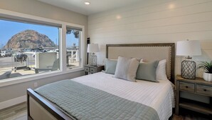 Un dormitorio con una cama grande, dos mesitas de noche con lámparas, vista a la montaña a través de una ventana y un balcón con barandilla.