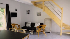 Ein gemütliches Wohnzimmer mit einer schwarzen Couch, zwei Korbstühlen, einem kleinen runden Tisch und einem Schrank. Im Hintergrund ist eine Holztreppe zu sehen.