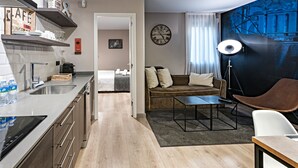 Ein modernes Hotelzimmer mit Küchenzeile, einer Couch, einem Esstisch und einem großen Fenster mit Vorhängen.