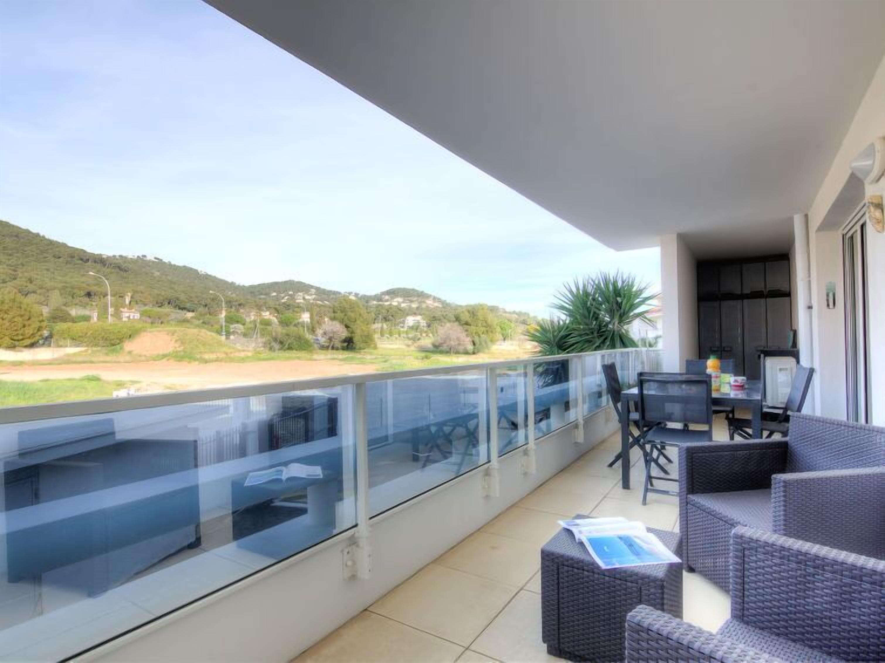 Un balcón con barandilla de vidrio, mobiliario exterior y vistas a un paisaje montañoso.