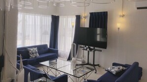 A modern living room with a blue sofa set, a glass coffee table, and a flat-screen TV mounted on the wall. - Thalassa Beach and Pool Villa (Yoron)