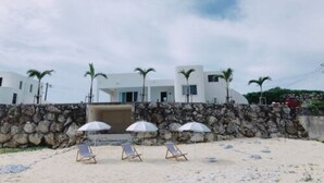 A beachside property with a stone wall, palm trees, and beach chairs with umbrellas. - Thalassa Beach and Pool Villa (Yoron)