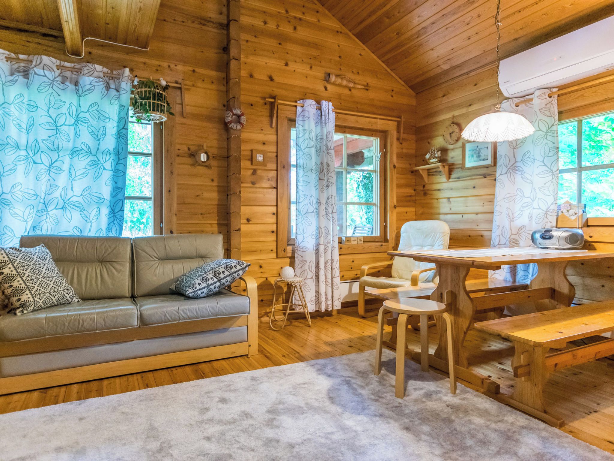 A cozy cabin interior with a wooden floor, a sofa with patterned cushions, a dining table with benches, and a window with a view of greenery.