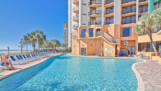 A swimming pool surrounded by a multi-story building with balconies, a clear sky, and palm trees. - Beautiful Oceanfront, Full Kitchen, Pet Friendly (Myrtle Beach)