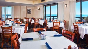 A hotel dining area with tables and chairs, large windows with curtains, and a view of the ocean. - Beautiful Oceanfront, Full Kitchen, Pet Friendly (Myrtle Beach)