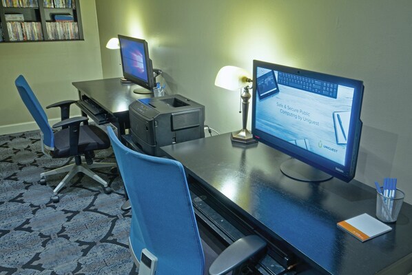 A hotel room with two monitors, a desk, a chair, a lamp, and a bookshelf with DVDs.
