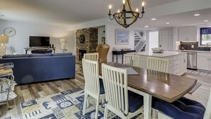 A modern living room with a blue sofa, a dining table with white chairs, a chandelier, a fireplace, and a kitchen in the background.