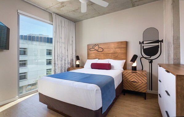 A hotel room with a bed, a nightstand with lamps, a dresser, and a view of buildings through the window. - DOWNTOWN Austin with ROOFTOP Pool and BBQ Area, Fire Pit & a Fitness Center (Austin)