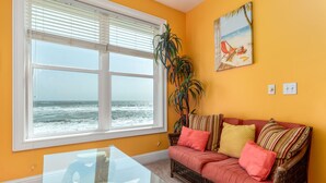 A living room with a large window overlooking the ocean, a glass coffee table, a wicker sofa with colorful cushions, and a tropical-themed painting on the wall. - Livin the Dream - Beachfront, 6/6, POOL, Pet Friendly, Sleeps 15 (Dauphin Island)