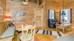 A cozy wooden cabin interior with a dining area, a sofa, a chair, and a small table with a lamp.