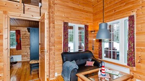 A wooden cabin interior with a black leather sofa, a glass coffee table, and a large window with curtains.