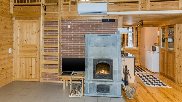 A wooden cabin interior with a stone fireplace, wooden walls, a loft area, and a kitchen with a microwave.